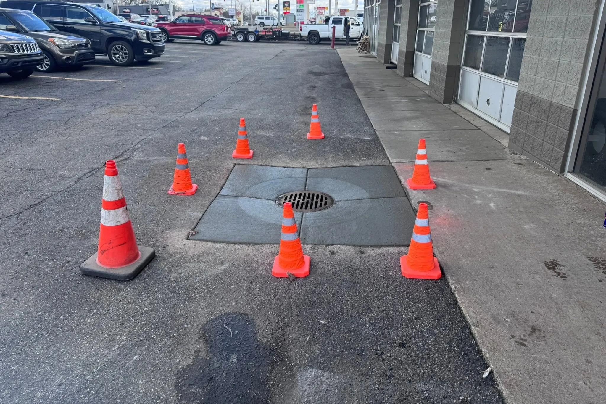 A new concrete pad finished flush around a storm drain in a commercial parking lot, coned off while it cures.