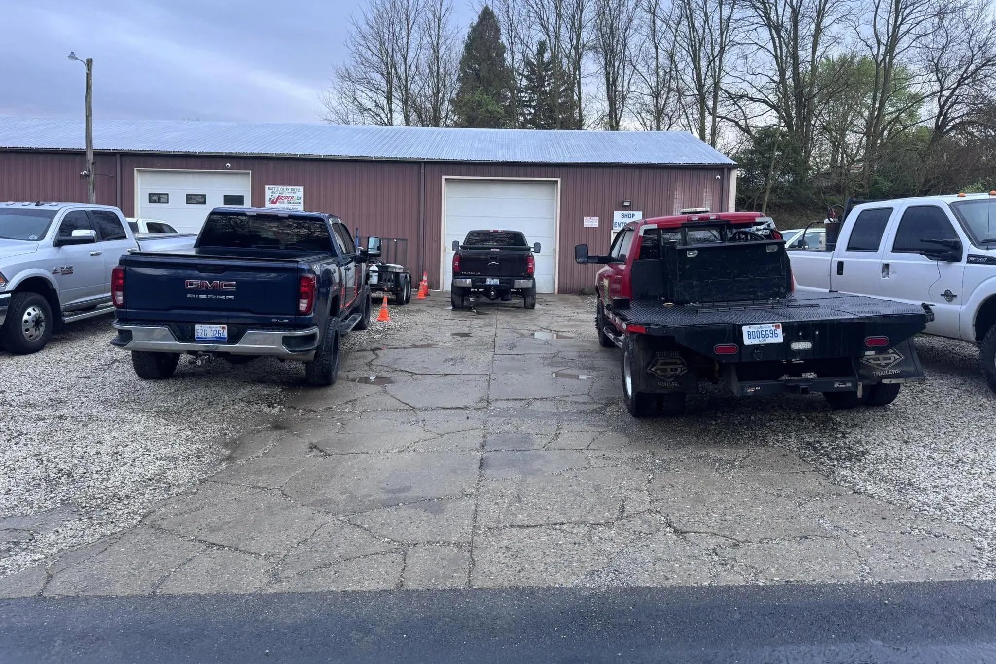 The approach to a commercial shop door before the work: broken asphalt and gravel holding puddles, with work trucks parked on either side.